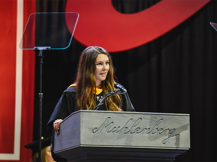 A college student with long brown hair wears a graduation robe and speaks at a podium engraved 