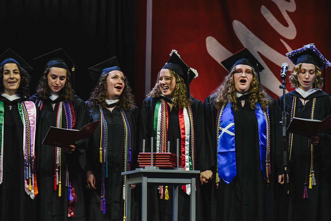 Six college graduates in commencement regalia sing and smile and hold hands on a stage